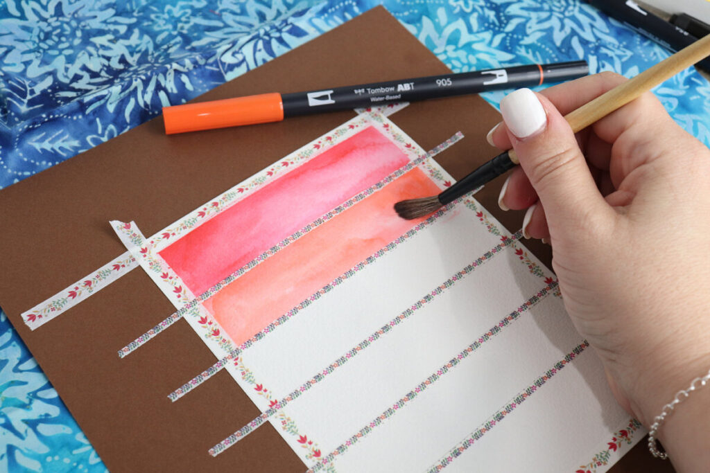 Image contains Amy’s hand holding a paintbrush and filling in sections of watercolor paper with colored ink. An orange marker sits nearby.