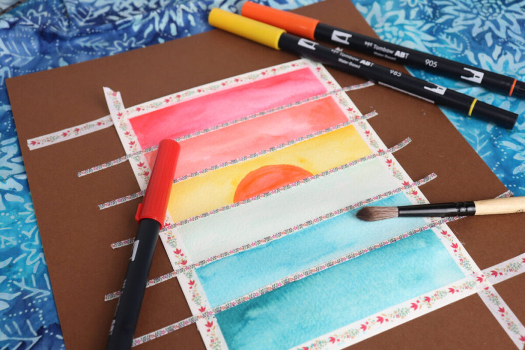 Image contains a piece of watercolor paper taped to a large piece of cardstock with washi tape. The sections are filled in with watercolor ink to create a sunset. A paintbrush and colored markers sit nearby.