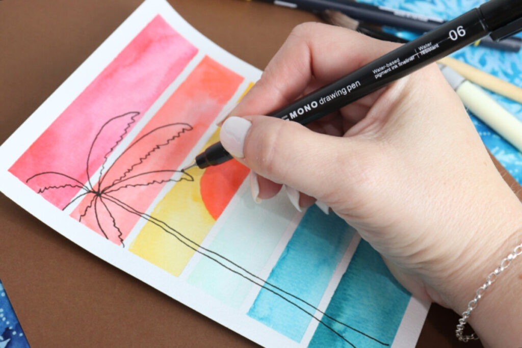 Image contains Amy’s hand holding a MONO Drawing Pen and sketching the silhouette of a palm tree onto a watercolor sunset postcard.