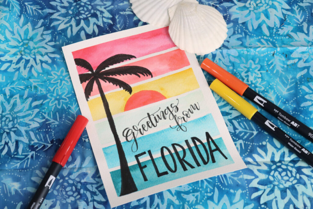 Image contains a colorful watercolor sunset postcard with a palm tree silhouette and the words “greetings from Florida.” Two scallop shells sit nearby along with three markers.