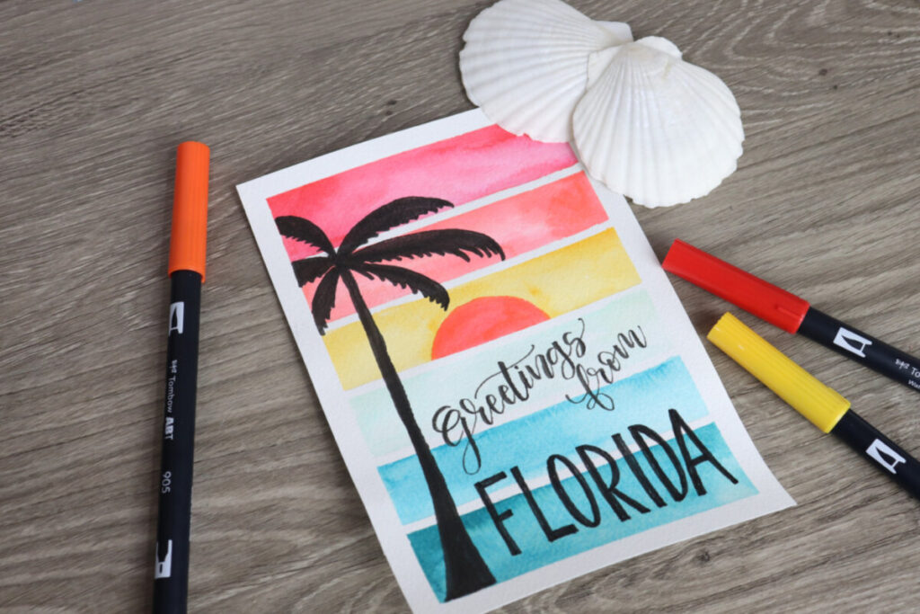 Image contains a watercolor sunset postcard with a palm tree silhouette and the words, “greetings from Florida” on a wooden desktop. Two scallop shells and three markers sit nearby.