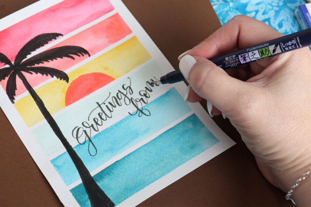 Image contains Amy’s hand holding a fudenosuke marker and lettering “greetings from” onto a painted postcard.