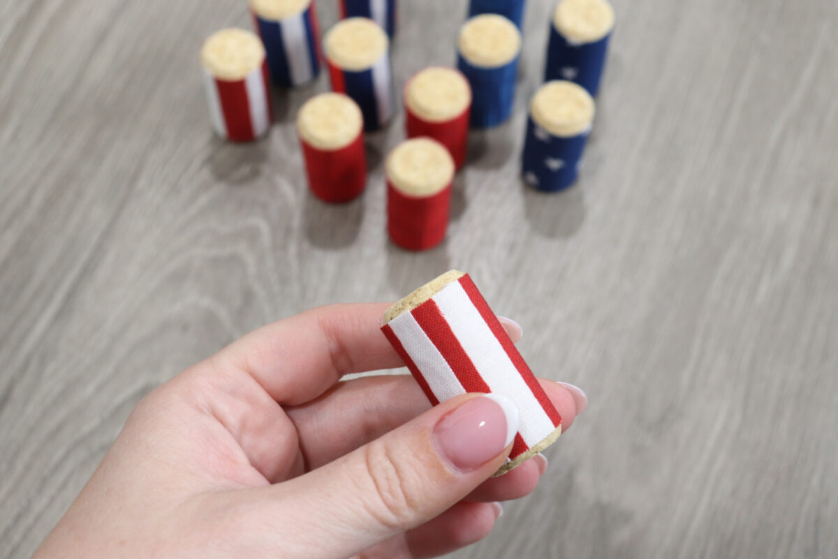 Image contains Amy’s hand holding a cork wrapped with a strip of red and white striped fabric. 11 other fabric wrapped corks sit on a wooden table in the background.