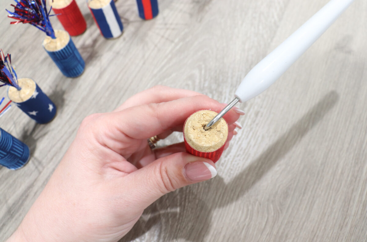 Image shows Amy’s hands using a pointed tool to poke a hole in the top of a cork. Other cork firecrackers sit off to the side.