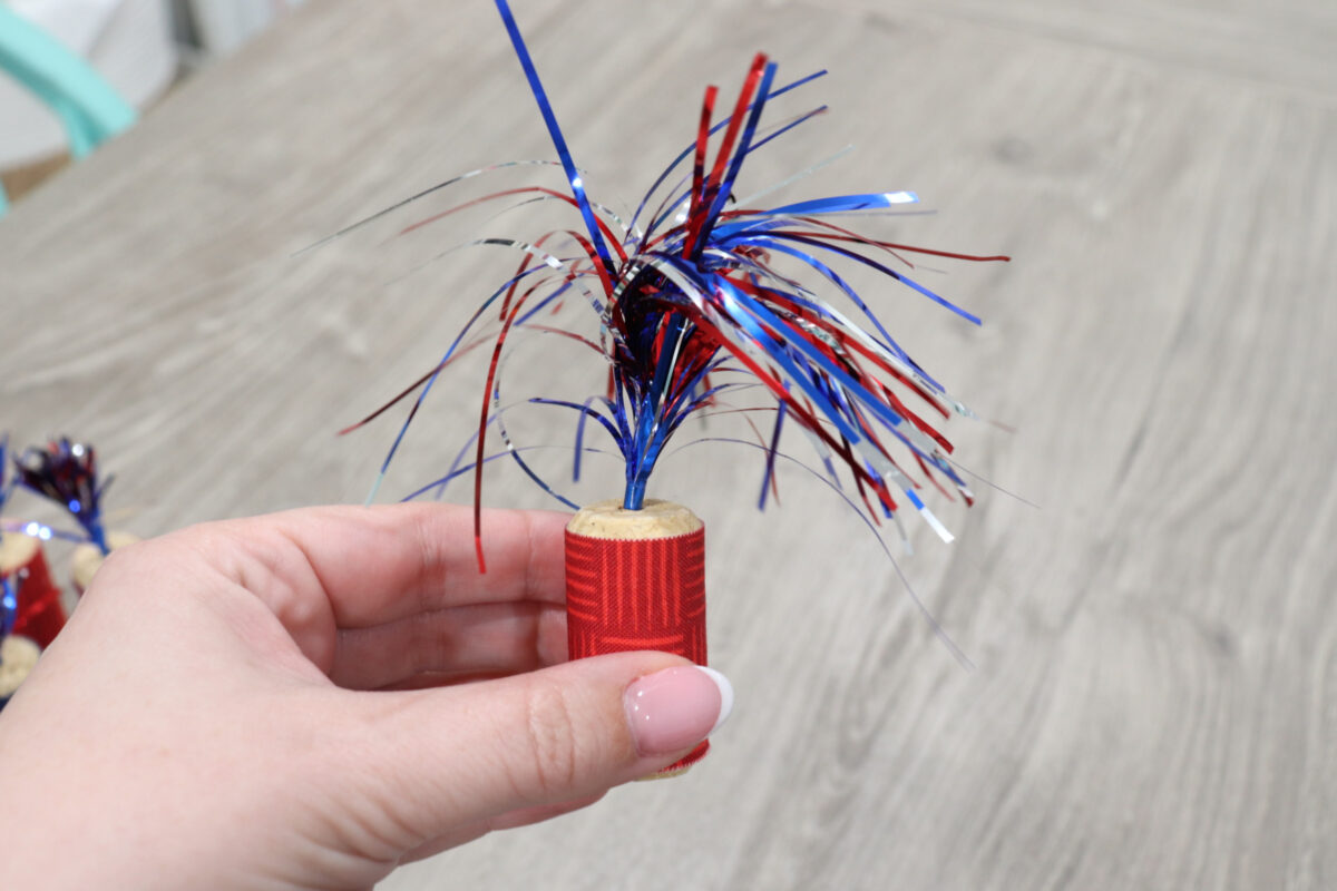 Image shows Amy’s hand holding a cork wrapped in red fabric with a patriotic cocktail pick inserted in the top.