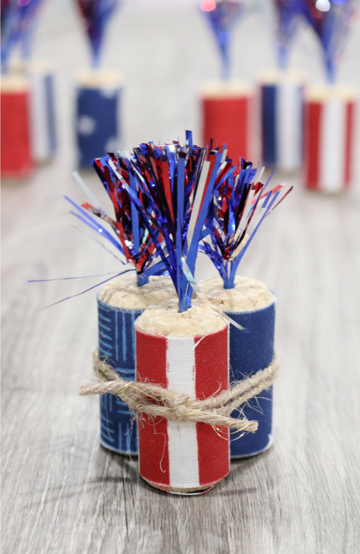 Image contains three cork firecracker decorations tied together with twine, sitting on a wooden desk. More firecrackers sit in the background, blurred.