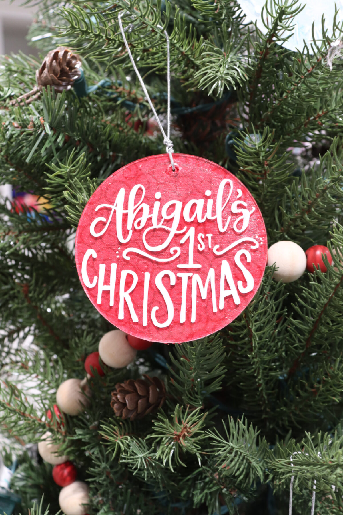 Image contains a round red ornament with the words, “Abigail’s 1st Christmas" in white lettering.