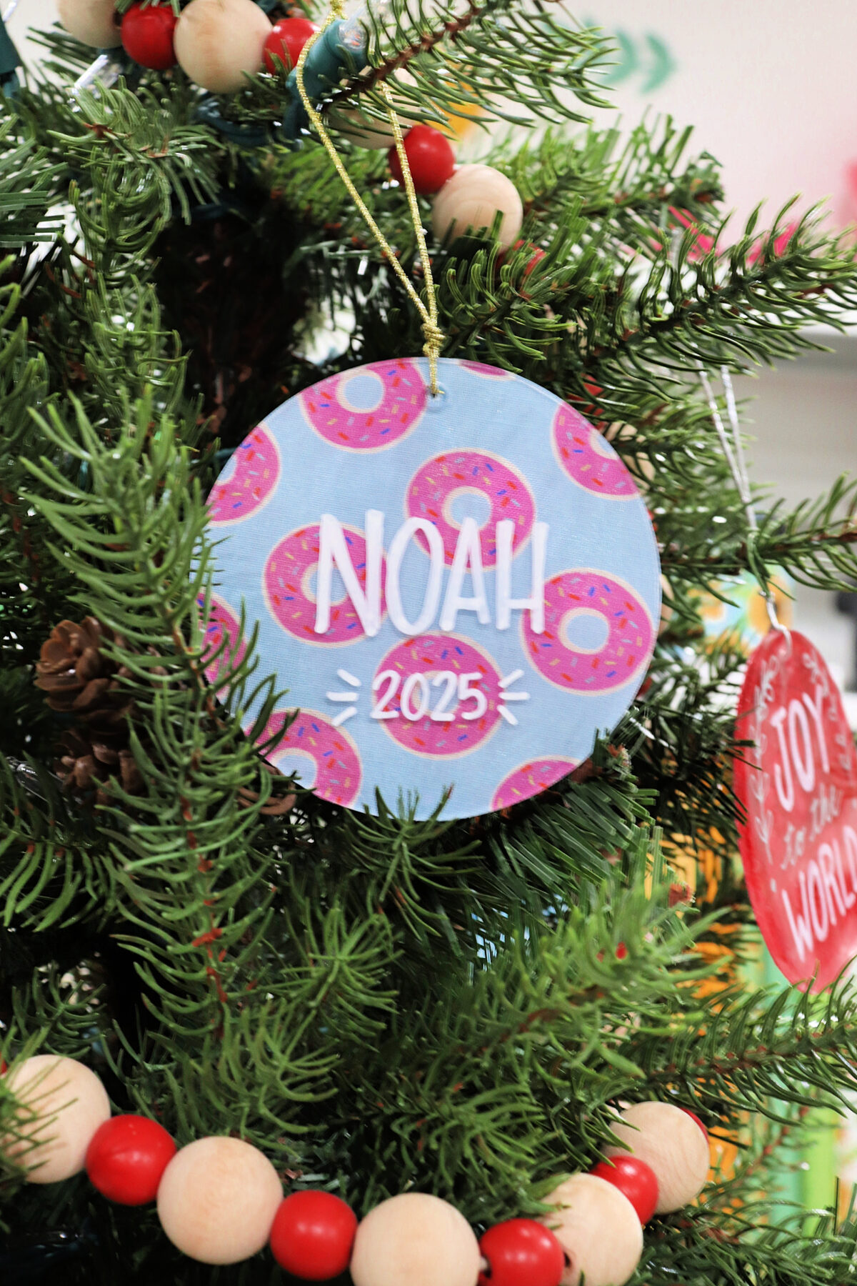Image contains a round blue ornament with pink sprinkled donuts, and “Noah 2025” written on it.