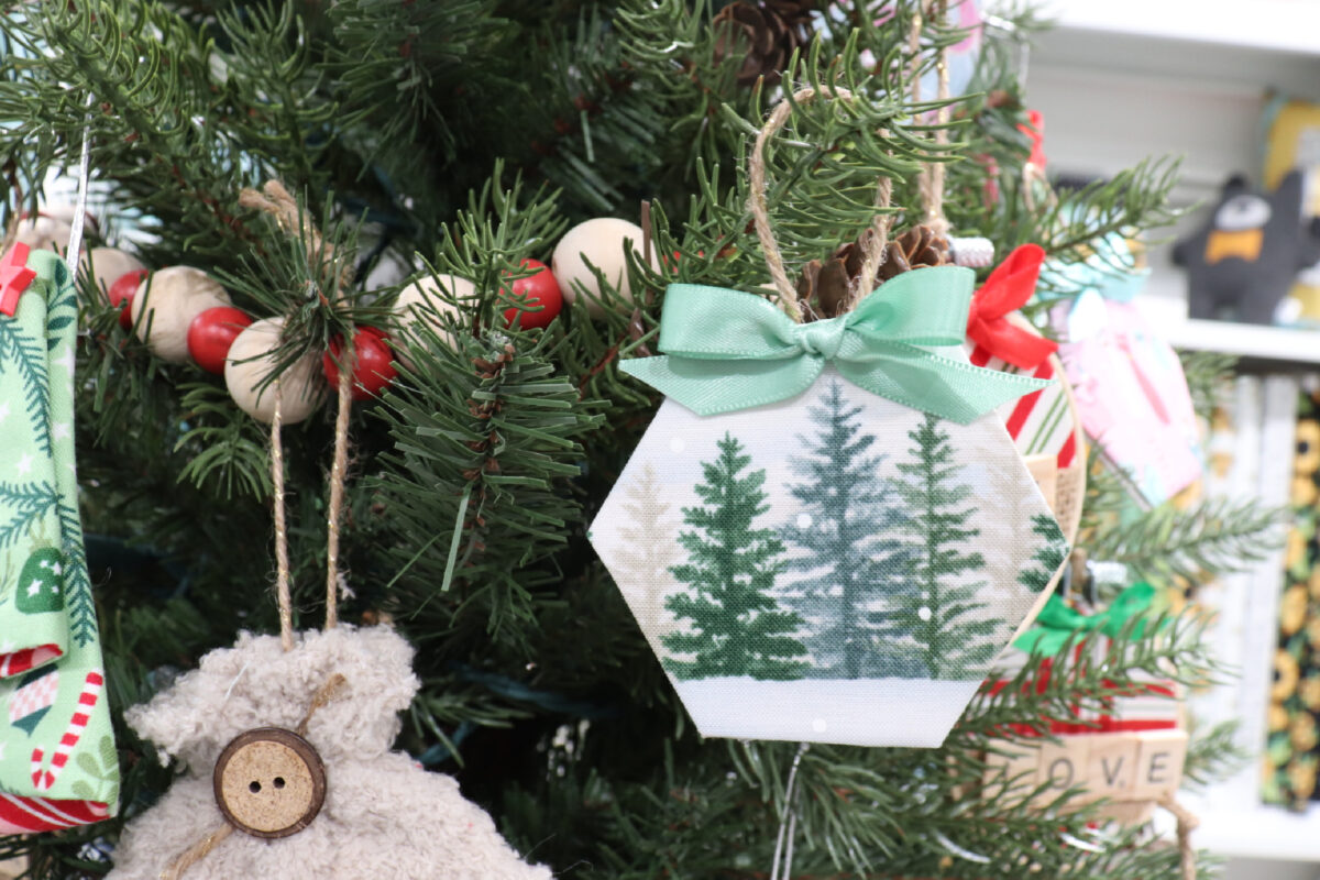 Image contains a fabric wrapped hexagon ornament featuring green and cream colored pine trees and a green bow.