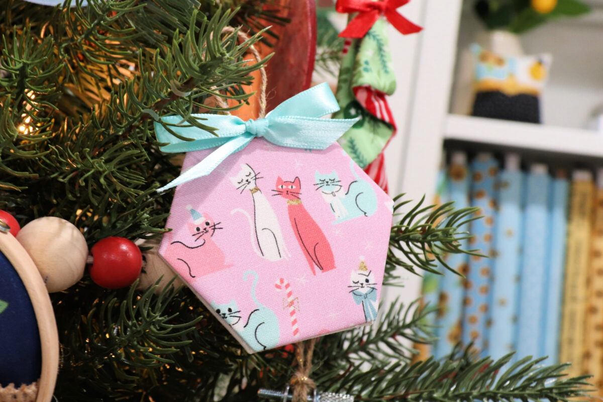 Image contains a pink fabric wrapped hexagon ornament with cartoon cats and a teal bow. It hangs on an artificial Christmas tree.