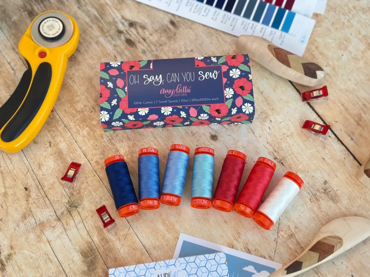 Image contains seven small spools of red, white, and blue thread, assorted red quilt clips, a yellow rotary cutter, and a blue thread box on a wooden table.