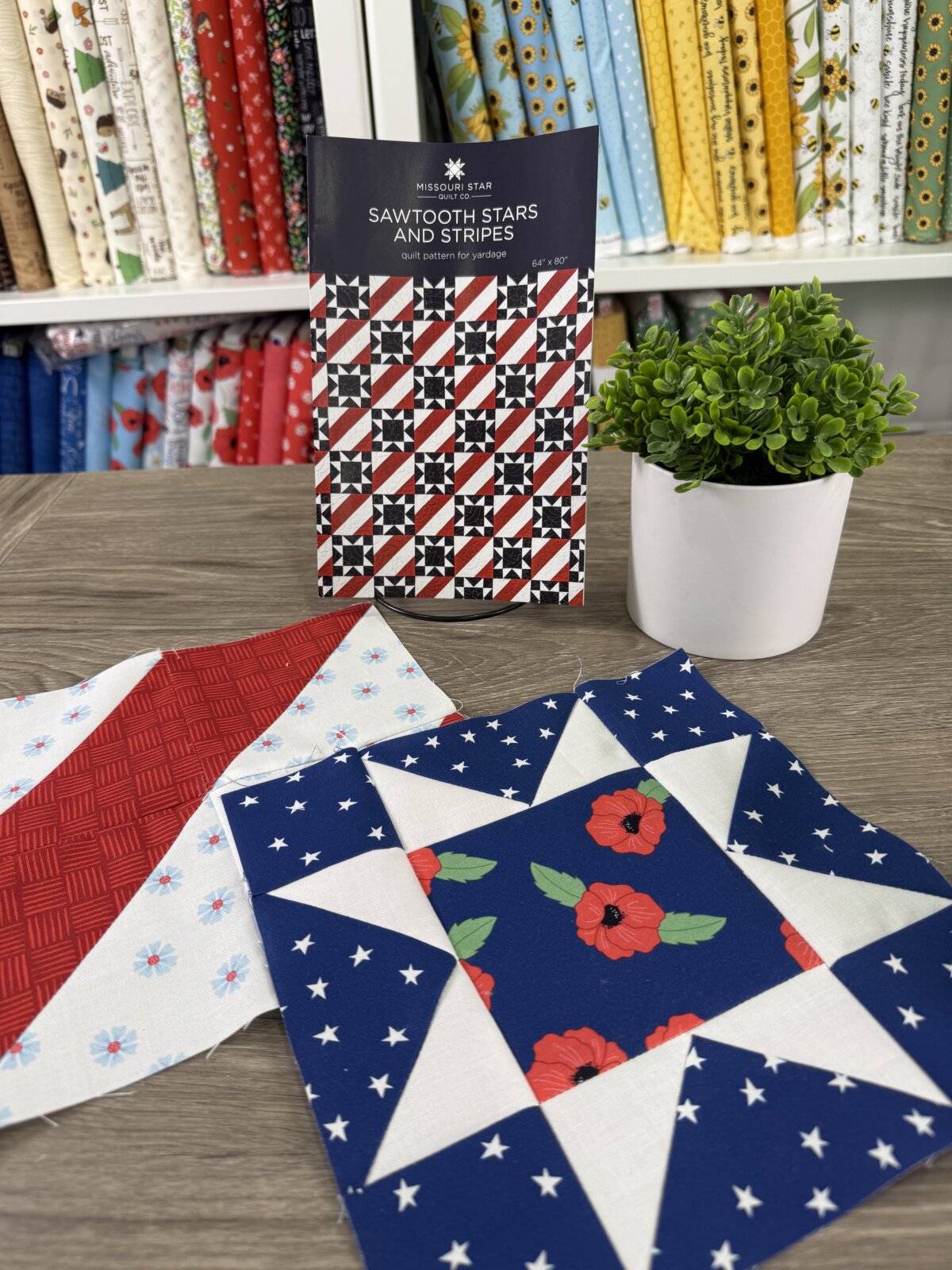Image contains two quilt blocks; one sawtooth star and one with stripes, laying in front of a quilt pattern and a small faux plant on a wooden table. Bolts of fabric are on shelves in the background.