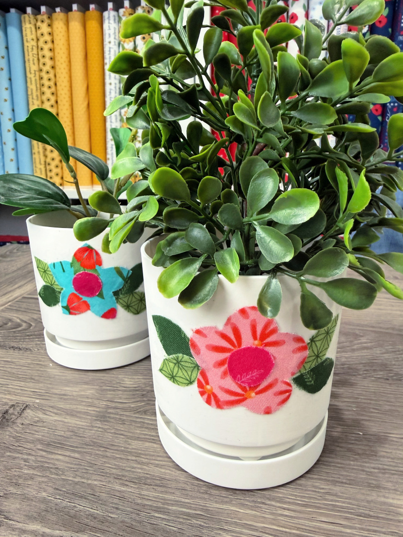 Image contains two faux plants in white plastic flowerpots decorated with fabric flowers.