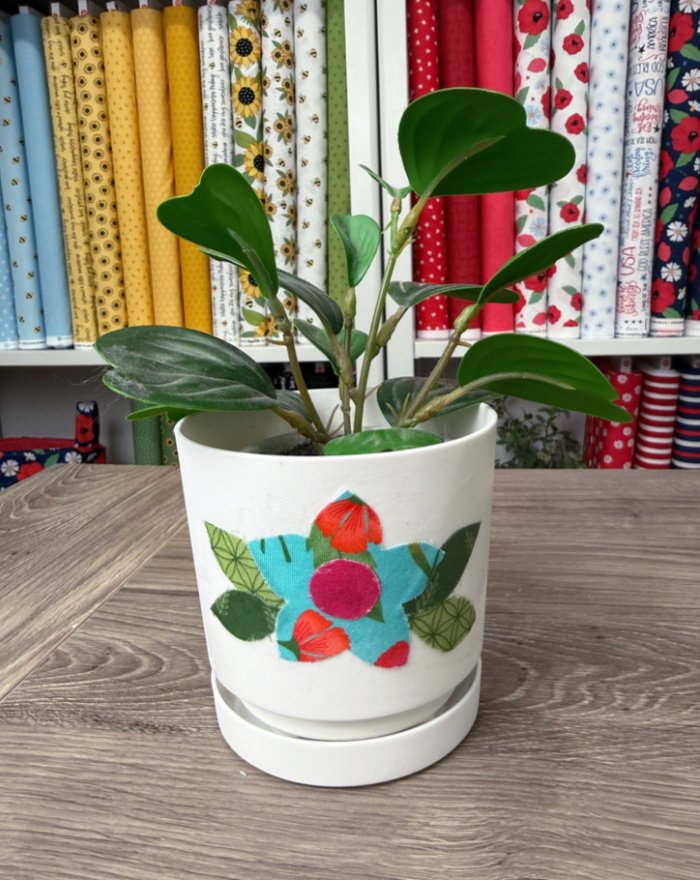 Image contains a faux plant in a white plastic flowerpot decorated with a fabric flower.