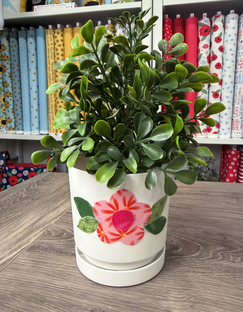 Image contains a faux plant in a white plastic flowerpot decorated with a fabric flower.