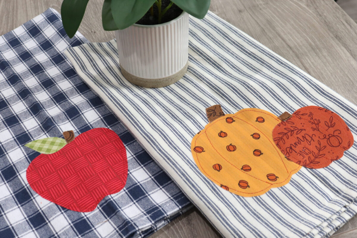Image contains two fall themed applique dish towels on a wooden desk with a faux plant.