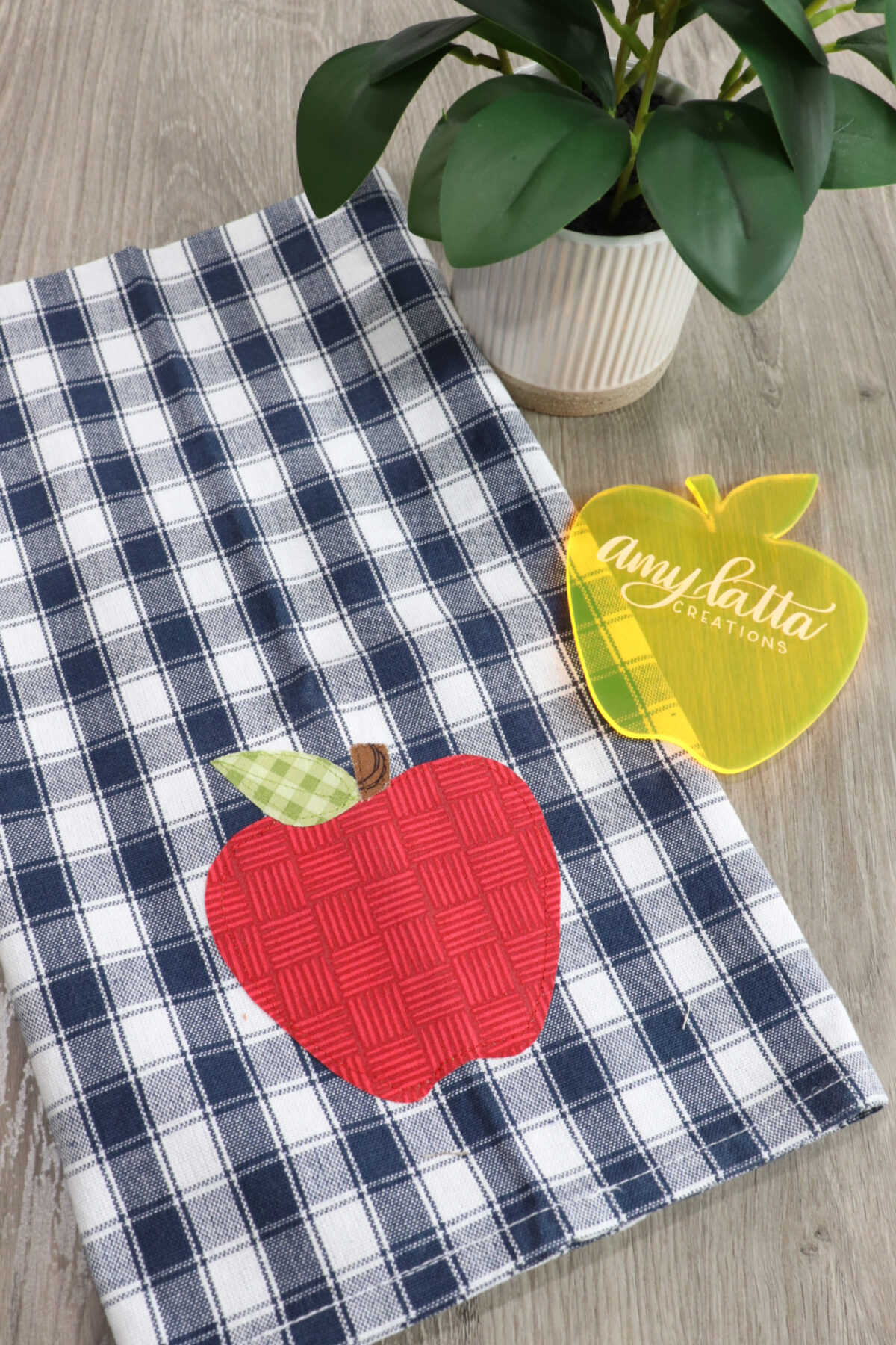 Image contains a blue and white checkered dish towel with a red apple applique. An apple template sits nearby on a wooden table top.