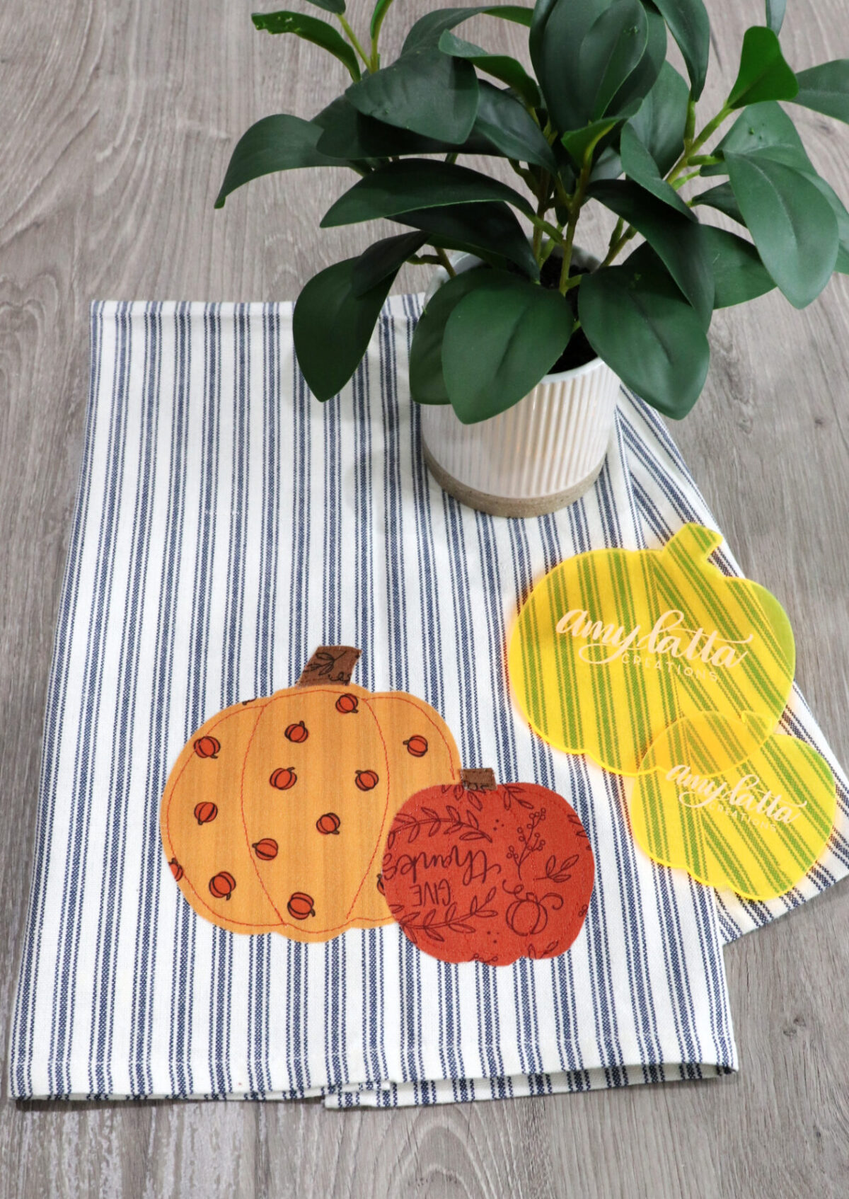 Image contains a blue and white striped dish towel with large and small pumpkins appliqued on the front. Pumpkin templates sit on top of the towel, as does a faux plant, all on a wooden desk.